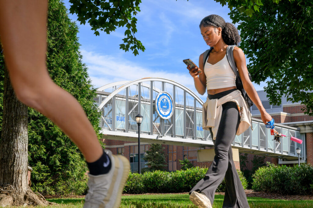 An MTSU student walks across campus while checking her phone near the pedestrian bridge featuring the Middle Tennessee State University seal on a sunny day.