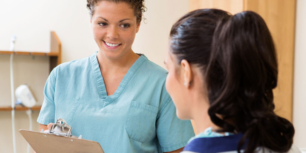 Medical assistant speaking with a patient while holding a clipboard in a healthcare setting.