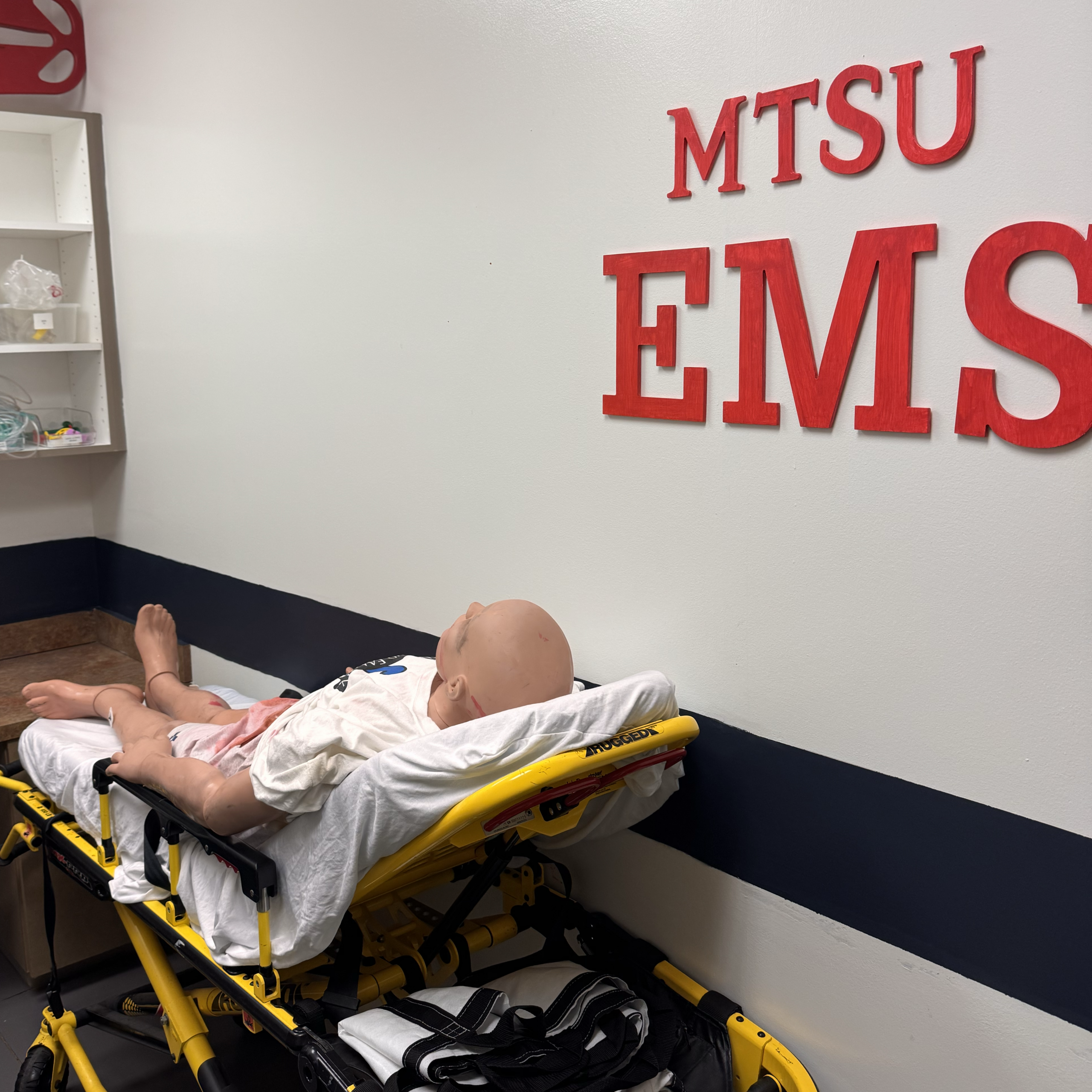 Patient simulation mannequin on an ambulance stretcher beneath MTSU EMS lettering.